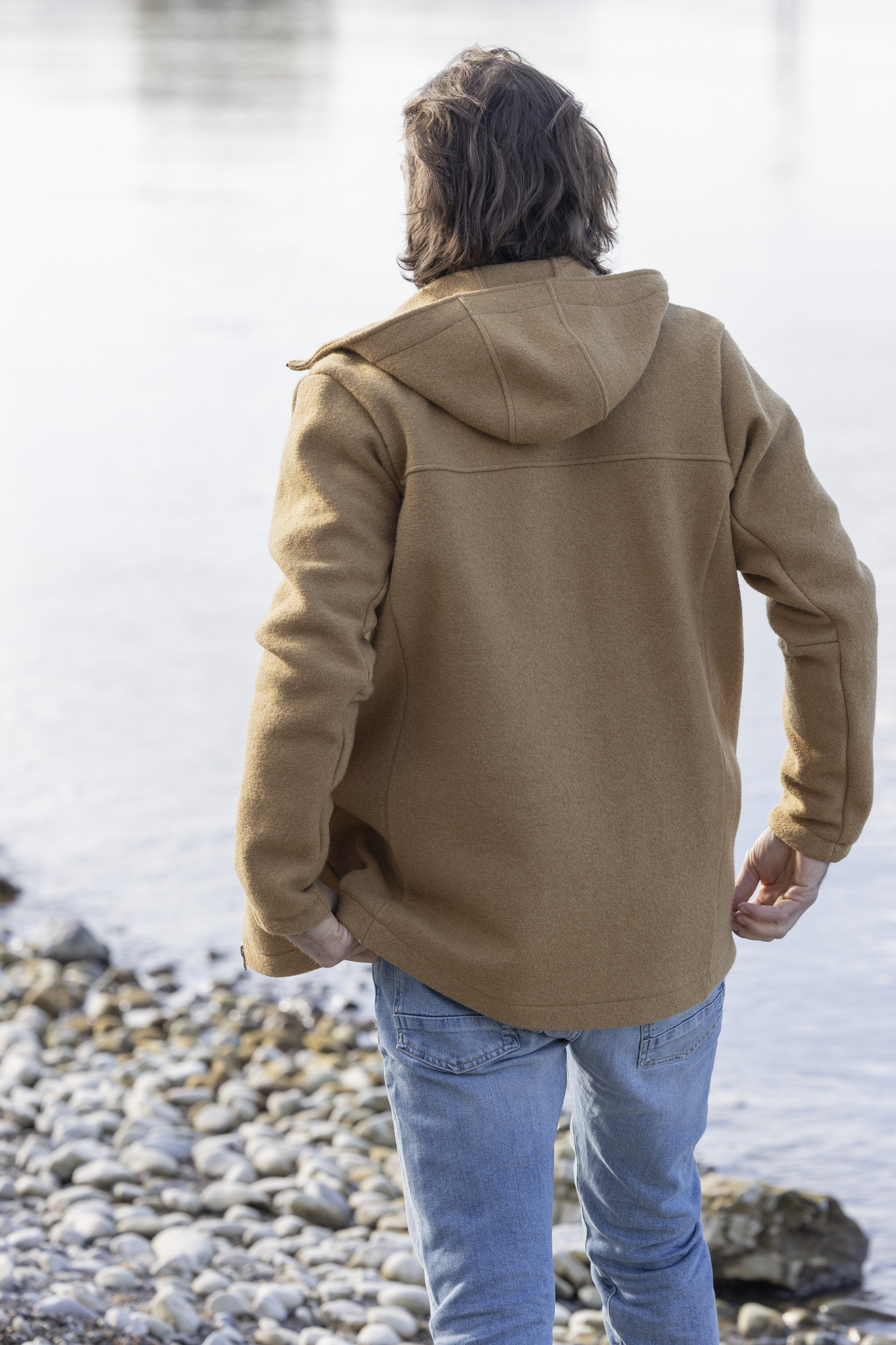 Rückansicht eines Mannes mit dunkelbraunen Haaren in disana Herren-Jacke Toffee und Jeanshose stehend an einem Seeufer