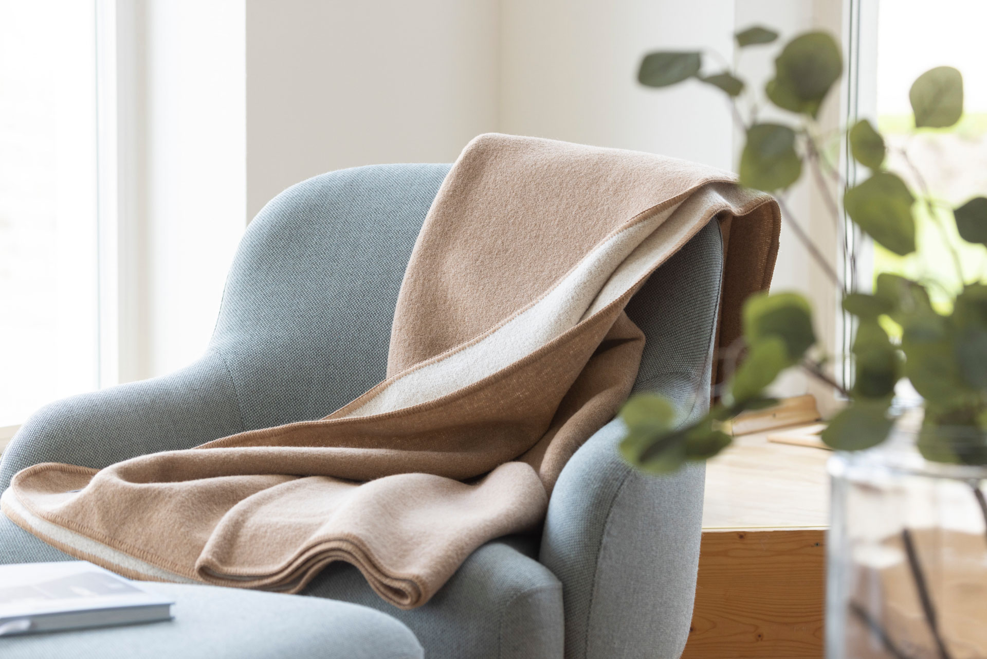 disana Doubleface-Decke in Karamell-Natur auf einem hellgrauen Sessel drapiert, im hellen Raum - Wollwalk aus Bio-Merinowolle