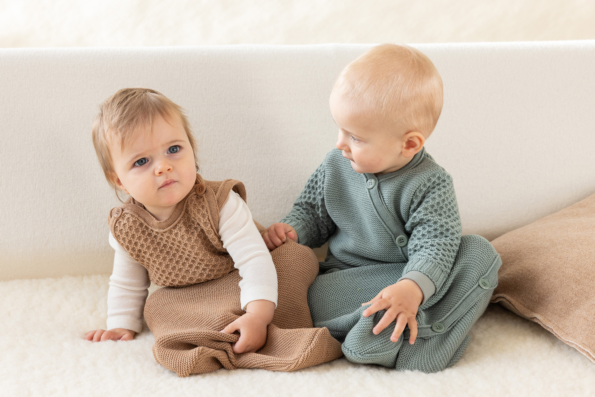 Zwei Babys in disana Träger-Schlafsack Karamell & Langarm-Schlafsack Jade sitzen auf einem Lammfell vor hellem Hintergrund