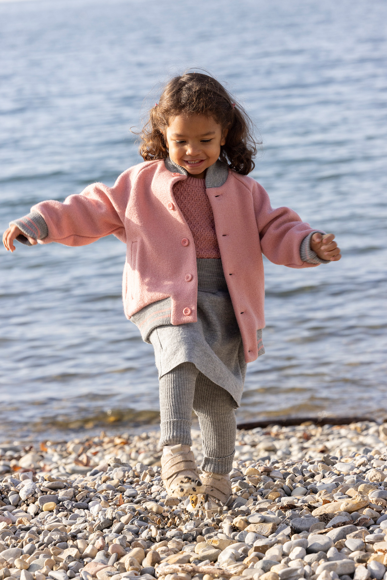 Kind mit hellbrauner Haut in disana College-Jacke & Aran-Pullover in Softpink, Walk-Rock & Strickleggings in Grau am Seeufer