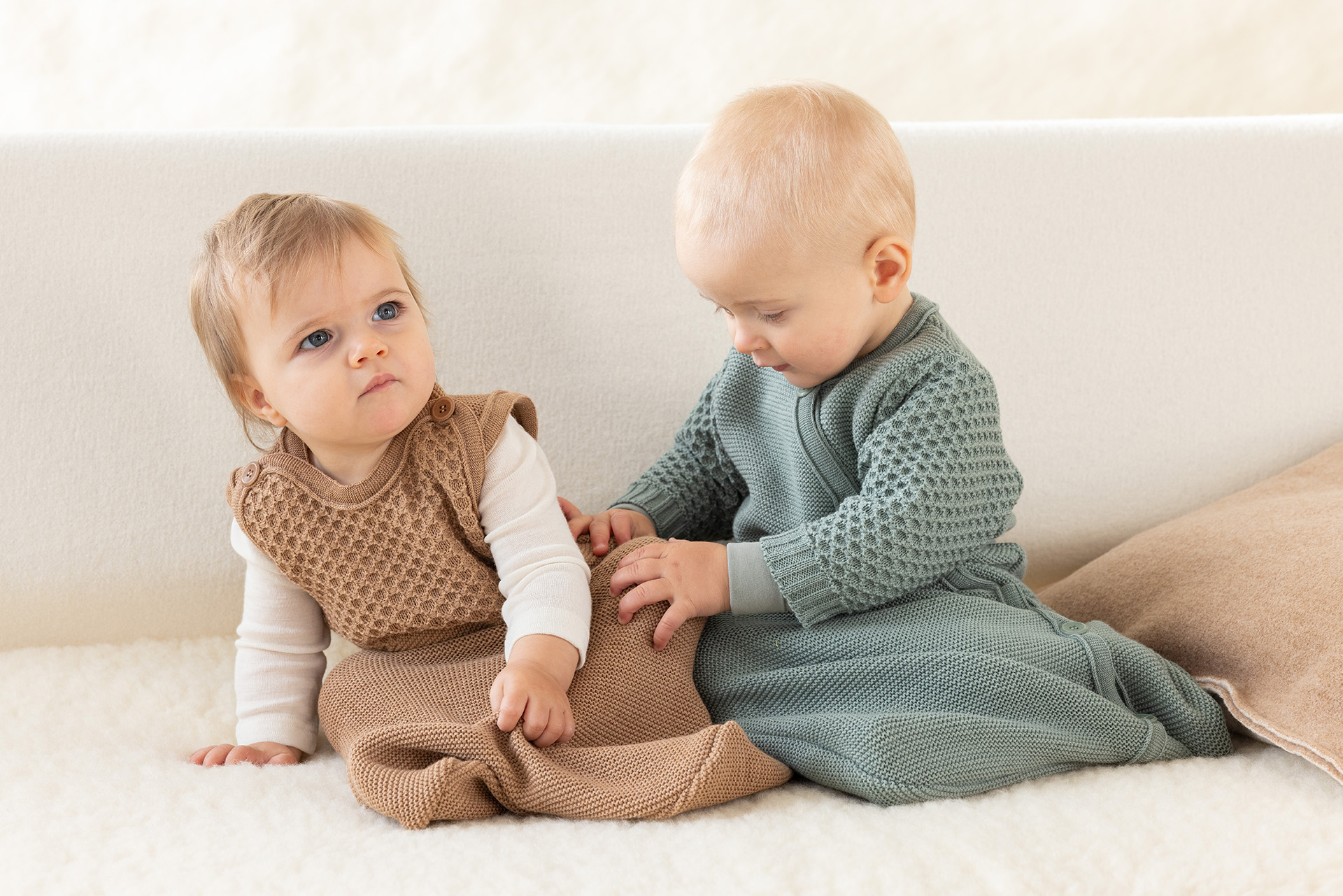 Zwei Babys in disana Träger-Schlafsack Karamell & Langarm-Schlafsack Jade sitzen auf einem Lammfell vor hellem Hintergrund