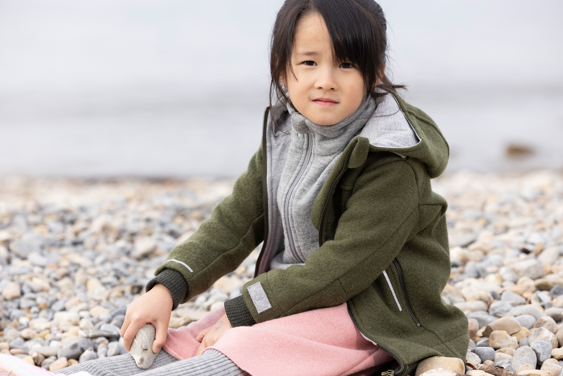 Asiatisches Mädchen in disana Outdoor-Jacke Oliv und Walk-Rock Softpink sitzt am Kieselstrand eines Sees & hält einen Stein