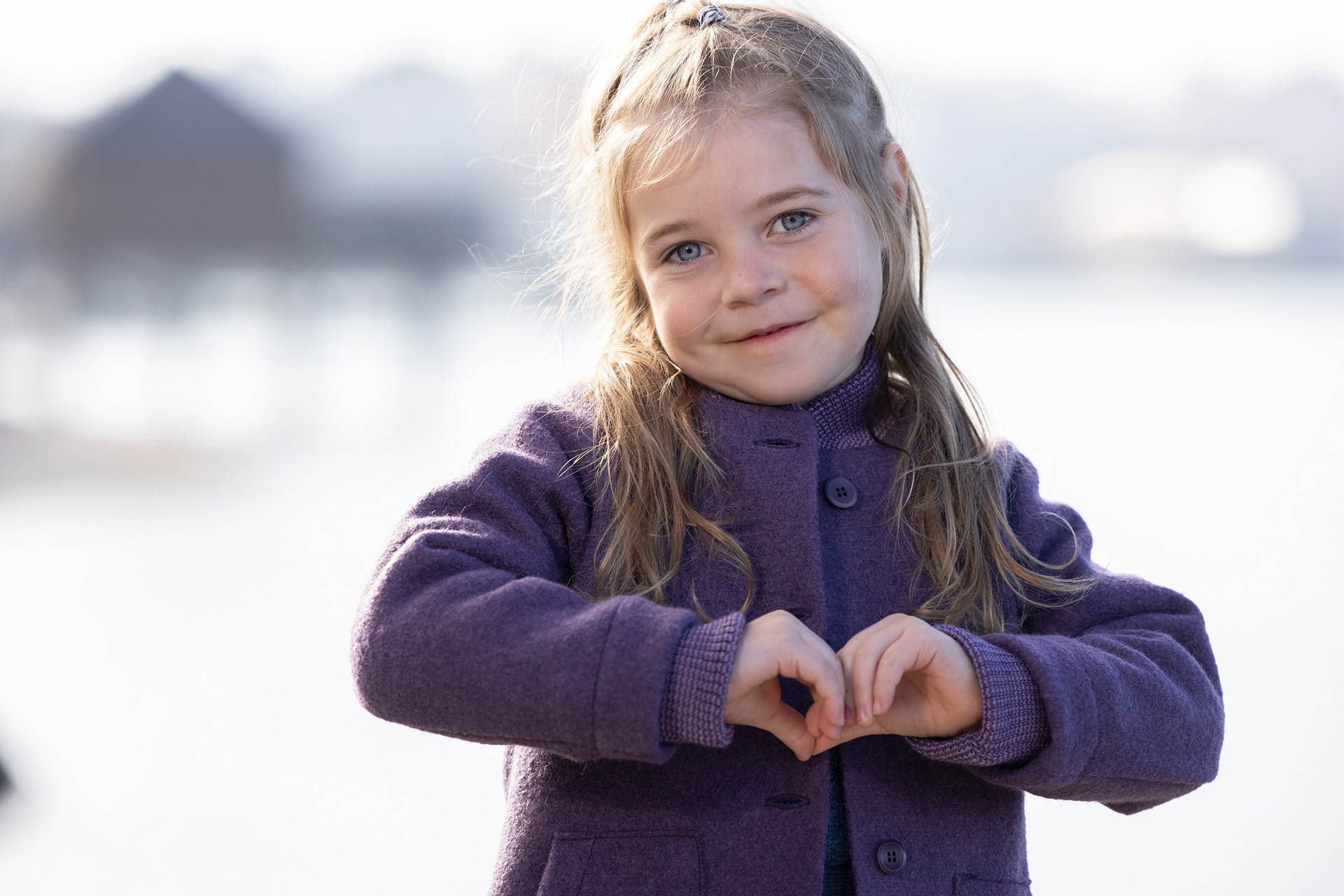 Ein blondes Mädchen im disana Walk-Mantel in Aubergine formt ein Handherz. Melierte Strickbündchen an Armen & Strickkragen 