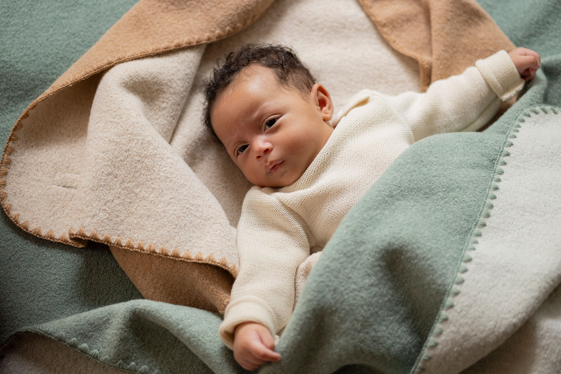 Baby mit hellbraunem Hautton eingekuschelt in disana Doubleface-Babydecken in den Farben Karamell-Natur und Jade-Natur