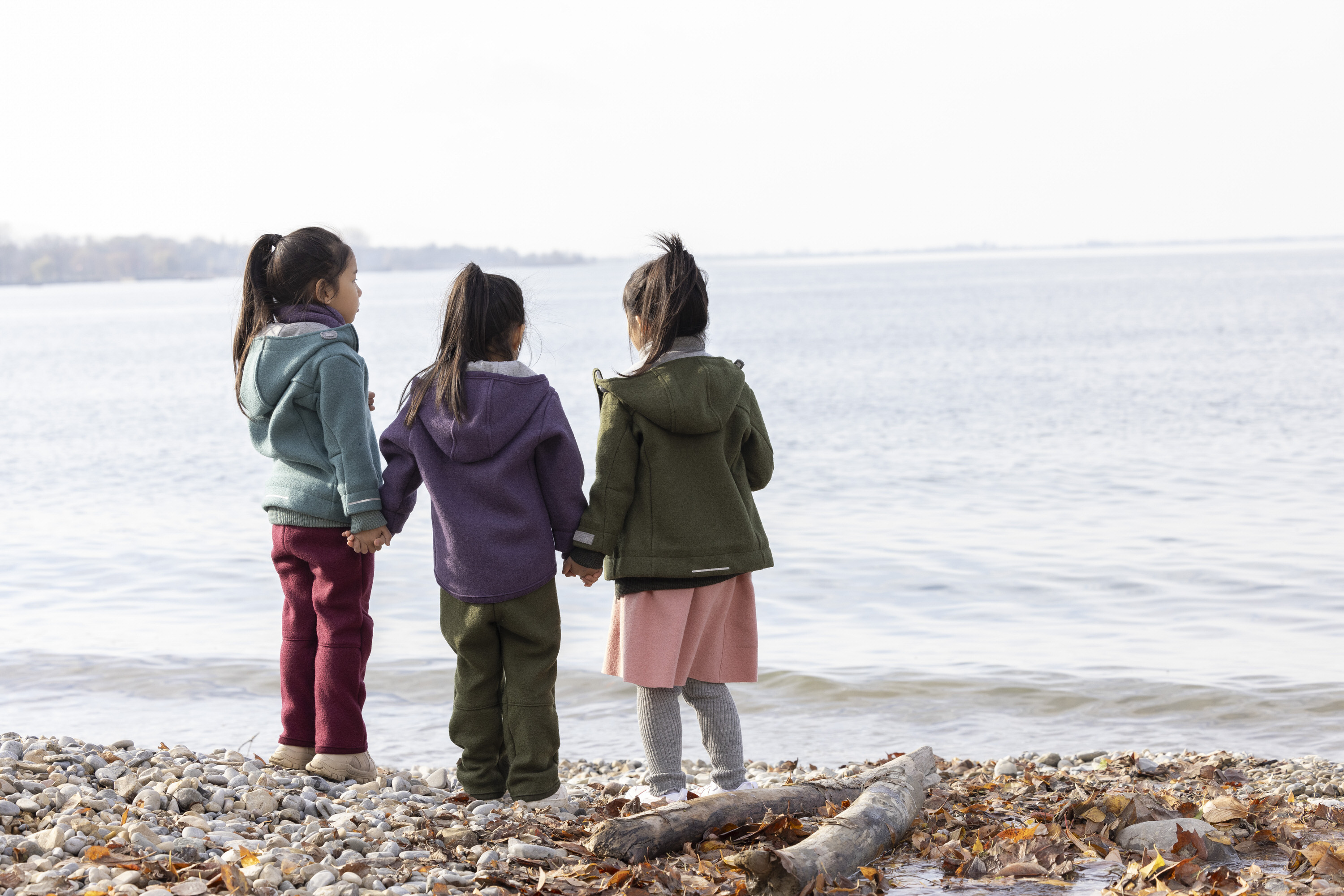 Rückansicht von drei Kindern in harmonierenden disana Wollwalk-Outfits, die gemeinsam auf das Wasser am Kieselstrand blicken