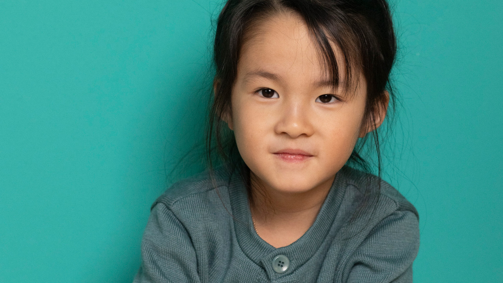 Asiatisches Mädchen in disana Strick-Jacke Eukalyptus mit großen Steinnußknöpfen aus Bio-Merinowolle vor grünem Hintergrund.