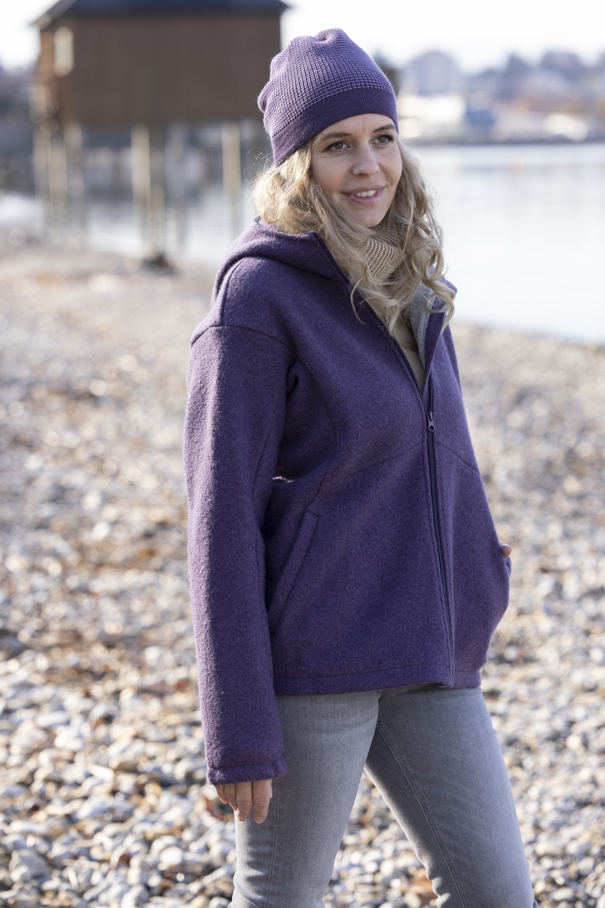 Lächelnde blonde Frau in disana Damen-Jacke Aubergine und passender Beanie Aubergine-Flieder am sonnigen Kieselstrand