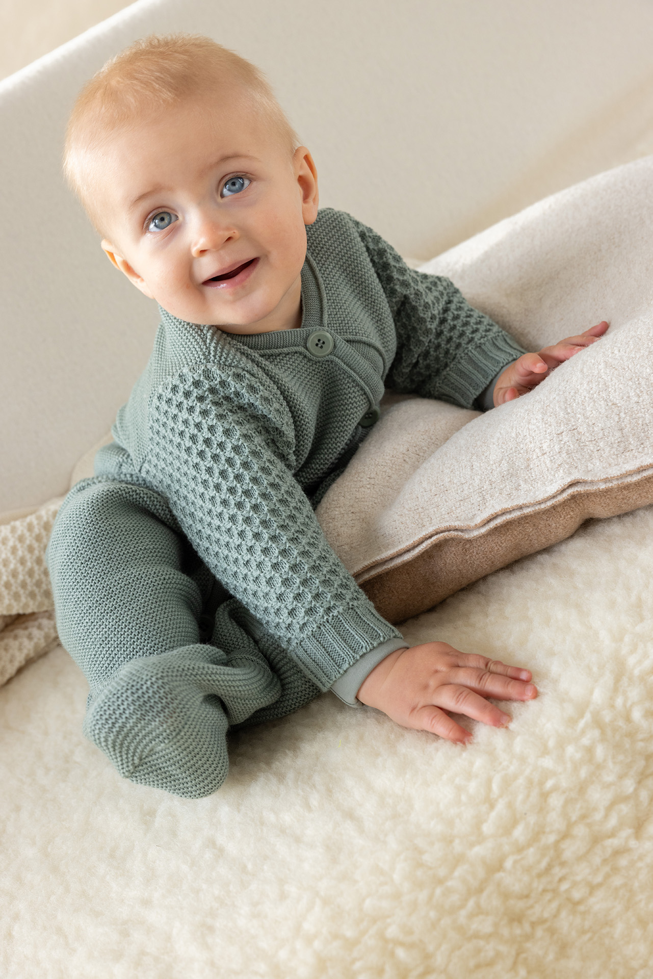 Blondes Baby mit blauen Augen in disana Strick-Overall in Jade aus Bio-Merinowolle auf einem flauschigen Lammfell