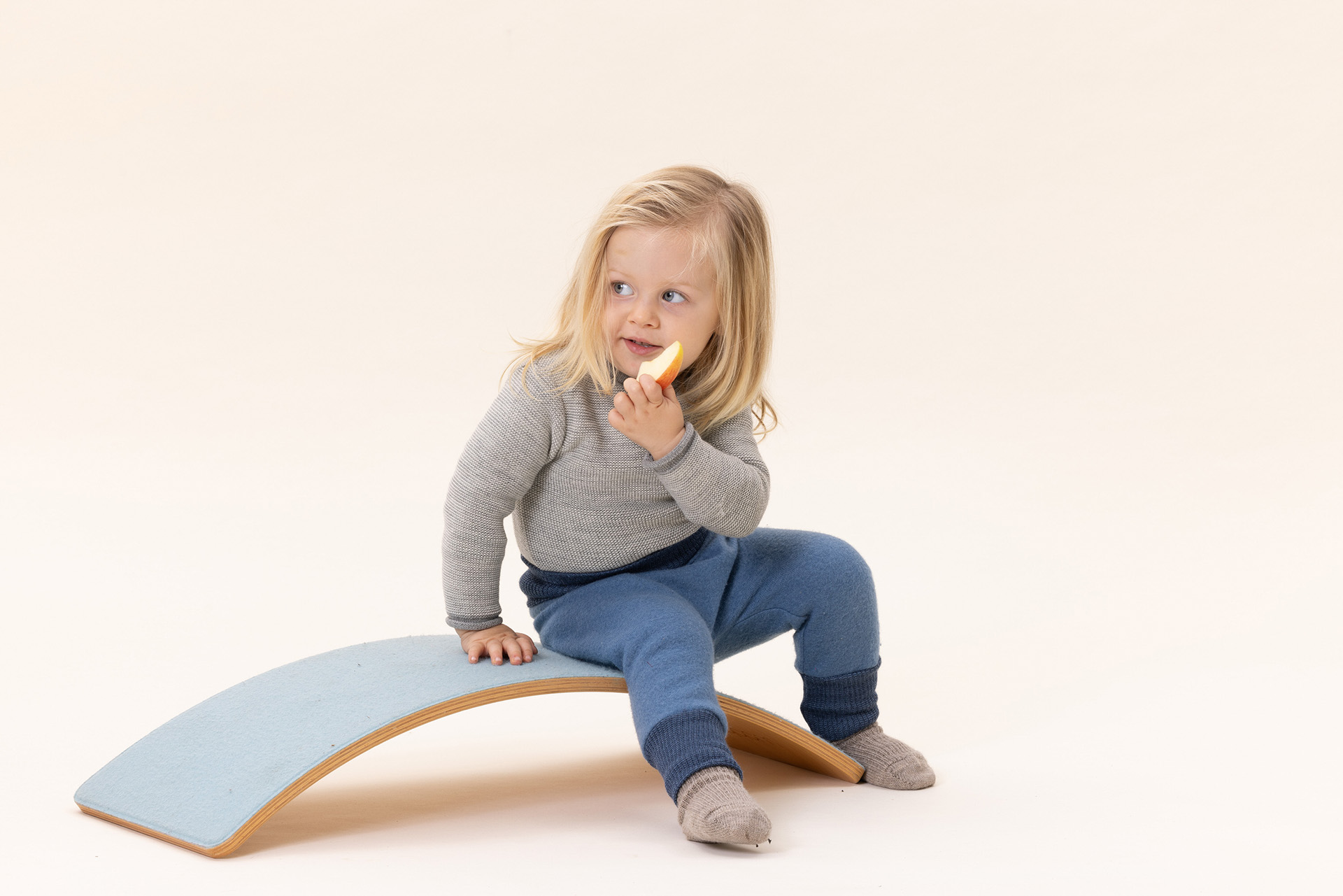 Blondes Kind auf Balanceboard in disana Pumphose Taubenblau, Melange-Pullover Grau-Natur, graue Socken vor beigem Hintergrund